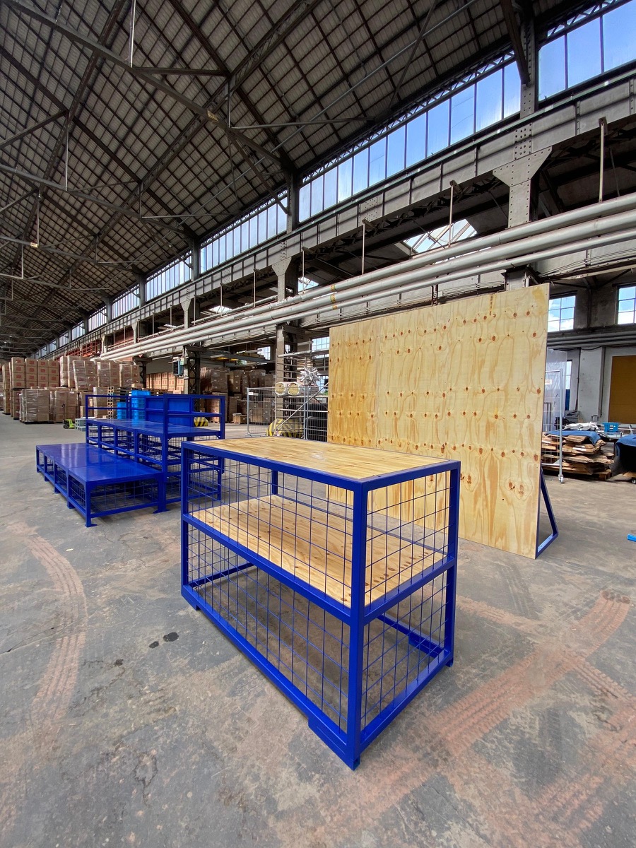 Blau lackierte Stahlmodule mit Gitterfüllung und Holzablagen in einer Halle